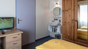 A hotel room with a bed, a wooden wardrobe, a bathroom with a sink and mirror, and a desk with a television.