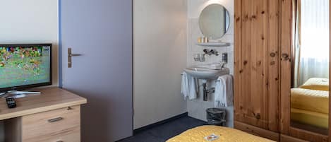 A hotel room with a bed, a wooden wardrobe, a bathroom with a sink and mirror, and a desk with a television.