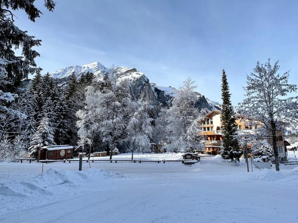 Paisagem coberta de neve com montanha ao fundo, um edifício com várias janelas e árvores carregadas de neve.