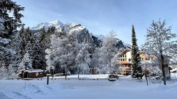 Paisagem coberta de neve com montanha ao fundo, um edifício com várias janelas e árvores carregadas de neve.