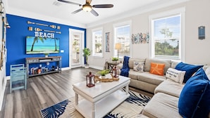 A living room with a blue accent wall, a ceiling fan, a television, a sofa, and a coffee table. - Beach Haven, Game Room, Arcades (Mexico Beach)