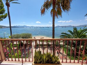 A balcony with a view of a calm sea, palm trees, and a distant mountain range. - Three bedroom apartment on the Pine Walk with superb views. WIFI and Air Con (Pollença)