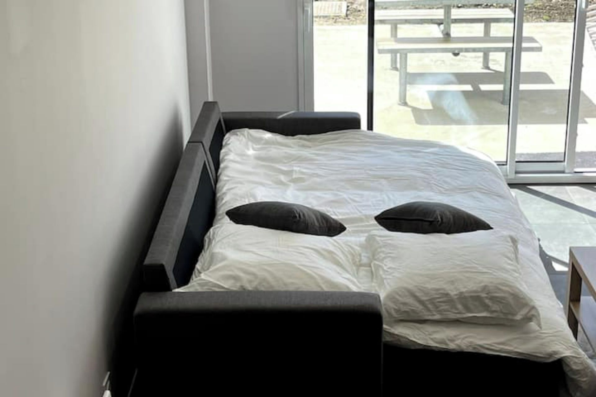 A bed with white bedding and two black pillows, a dark headboard, and a view of an outdoor patio through a large window.