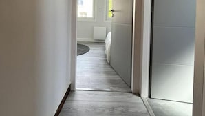 Un couloir étroit, doté d’un plancher en bois, mène à une chambre à coucher qui contient un lit et une fenêtre.