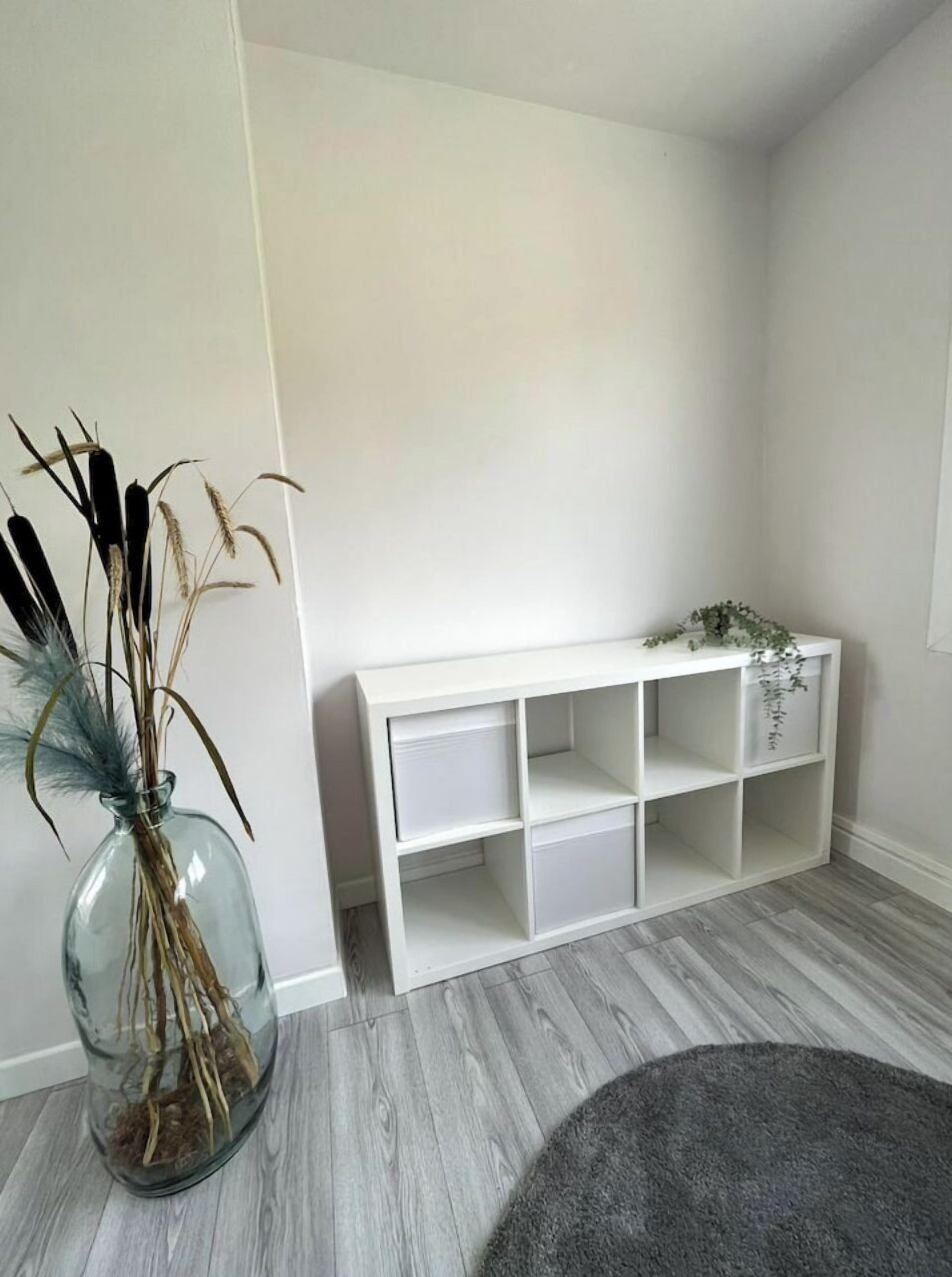 Une pièce avec une console blanche, un vase en verre contenant des plantes séchées et un tapis gris.