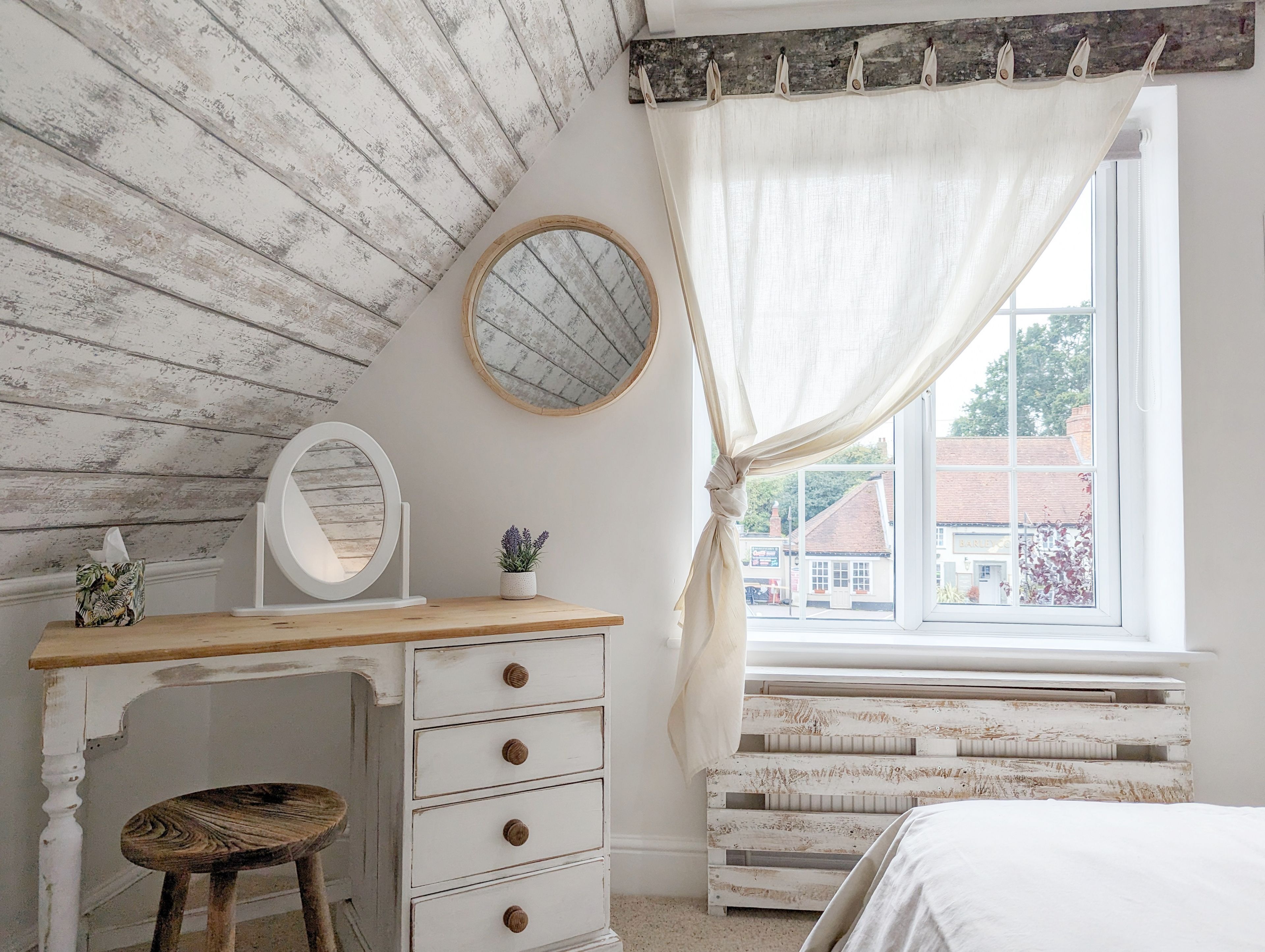 Un dormitorio con techo de madera, una mesita de noche con cajones, un espejo redondo, una ventana con cortinas y vistas a edificios.