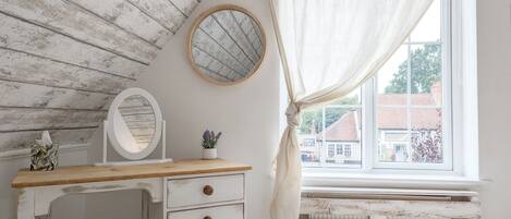 Un dormitorio con techo de madera, una mesita de noche con cajones, un espejo redondo, una ventana con cortinas y vistas a edificios.