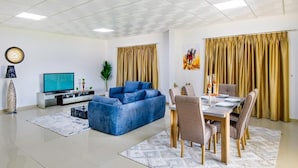 A modern living room with a blue sofa, a dining table with chairs, a TV, and a plant. - Nordic Gardens (Jannusan)