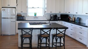 Une cuisine avec des meubles blancs, une cuisinière, un four à micro-ondes et un évier. On y trouve trois tabourets de bar et une fenêtre au-dessus de l’évier.
