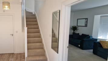 A staircase leading to a second floor, a living room with a dark blue sofa, a large mirror, and a potted plant.