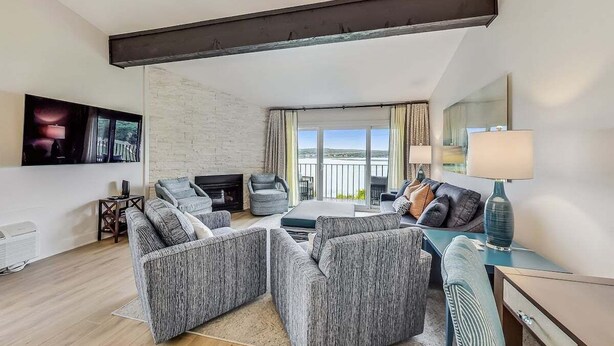 A modern living room with a fireplace, grey upholstered furniture, a TV, and a balcony with curtains. - Glass Vista: Modern 2BR Condo, Lake Views (Lake Geneva)