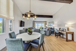 A modern living room with a round dining table, comfortable seating, a chandelier, and a large window.