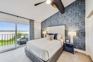 A bedroom with a large bed, two bedside tables with lamps, a patterned wallpaper, and a balcony with outdoor furniture.