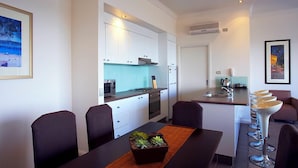 Cozinha moderna com armários brancos, forno embutido e micro-ondas. Há uma área de jantar com mesa e cadeiras, e uma pequena ilha com pia e banquetas.