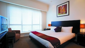 Quarto de hotel com cama, escrivaninha, cadeira, televisão e um quadro na parede.