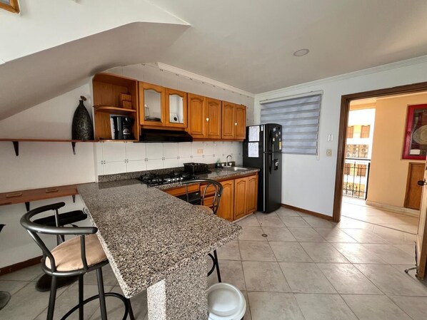 A kitchen with granite countertops, wooden cabinets, a black refrigerator, and a window with blinds. - Edificio El Peñón del Rodadero (Santa Marta)