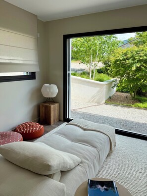 A bedroom with a large window, a bed with white bedding, a small wooden side table, and a red patterned ottoman.