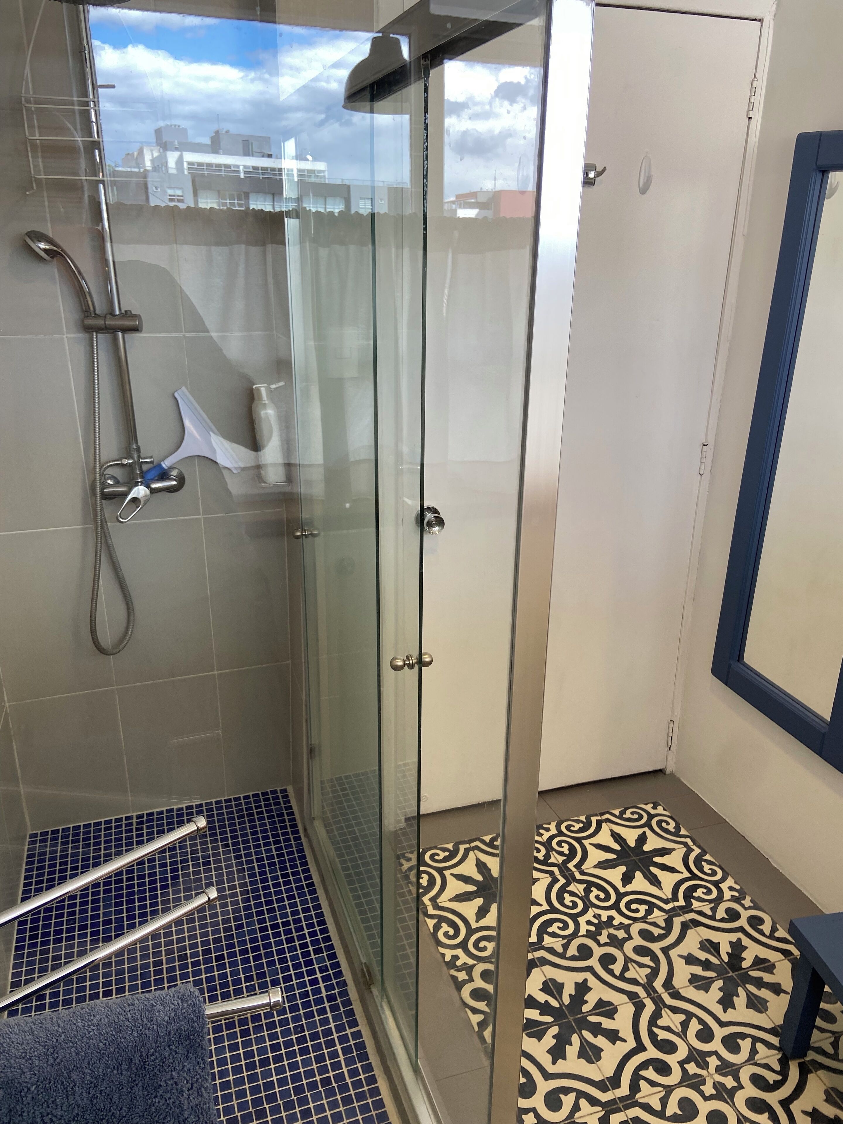 A glass-enclosed shower with a handheld showerhead and a handheld sprayer, a glass door, and a patterned floor.