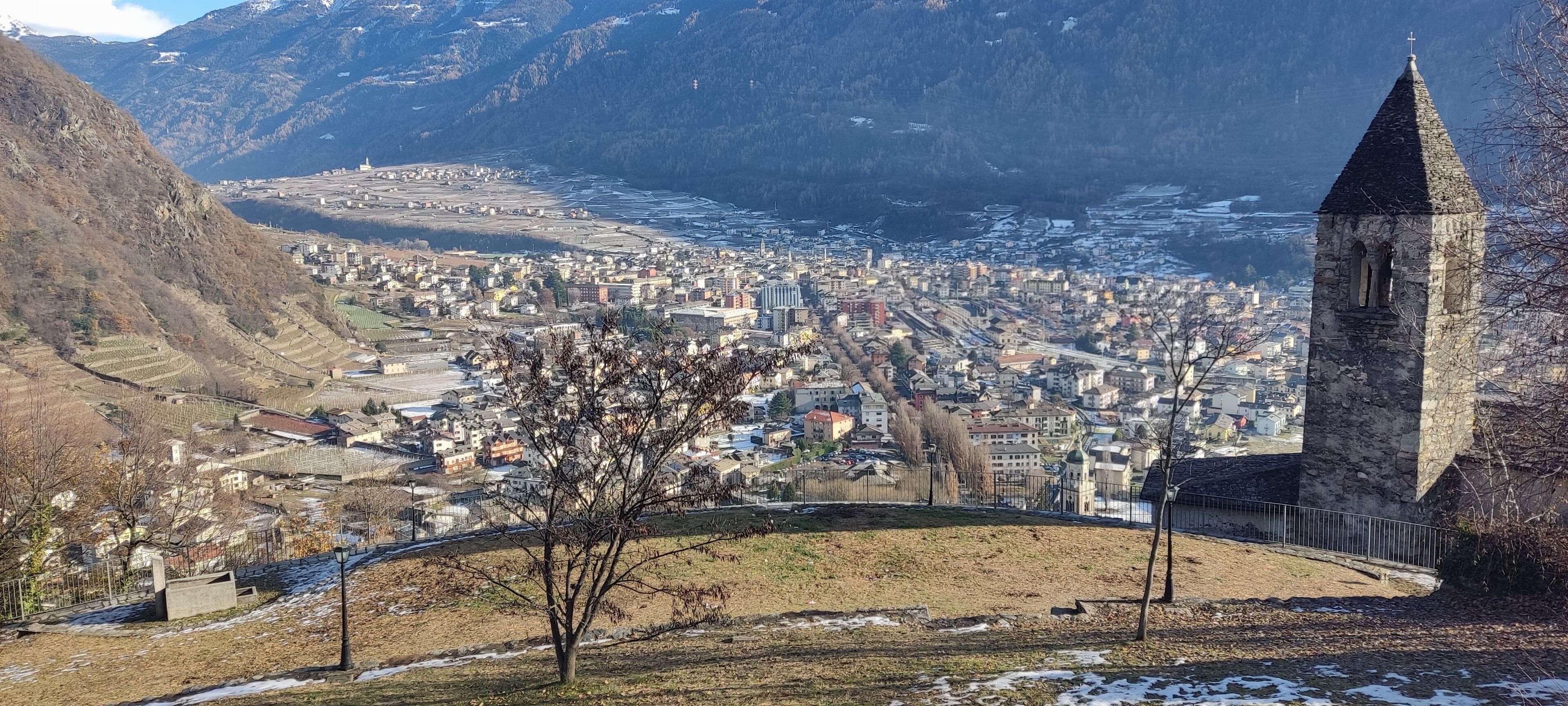 A town with a prominent stone tower, a river, and surrounding mountains.
