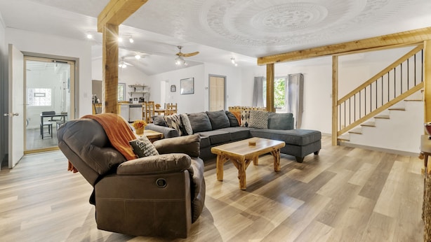 Amplia sala de estar con un sofá seccional grande, un sillón reclinable y una mesa de centro de madera. Se ve una escalera con barandillas de madera al fondo.