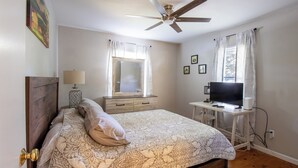 Ein Schlafzimmer mit einem Bett, einem Schreibtisch mit Fernseher, einem Deckenventilator und einem Fenster mit Vorhängen.