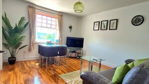 A spacious living room with a wooden floor, a sofa, a dining area with a table and chairs, a TV, and a large window with curtains.