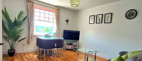 A spacious living room with a wooden floor, a sofa, a dining area with a table and chairs, a TV, and a large window with curtains.