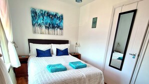 A bedroom with a large bed, wooden flooring, a ceiling light, a painting on the wall, and a door with a mirror.