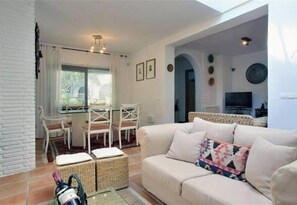 Uma sala de estar com um sofá bege, um espaço de refeições com cadeiras brancas e uma TV na parede.