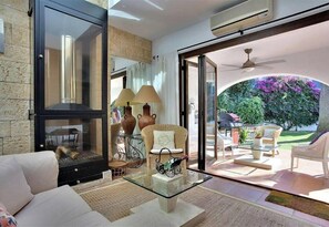 Uma sala de estar moderna com lareira de vidro, mobiliário de vime e vista para um jardim com flores roxas.