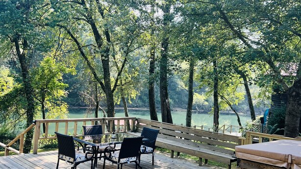 Une terrasse en bois, agréée de mobilier d’extérieur, qui offre une vue sur une étendue d’eau et est entourée d’arbres.