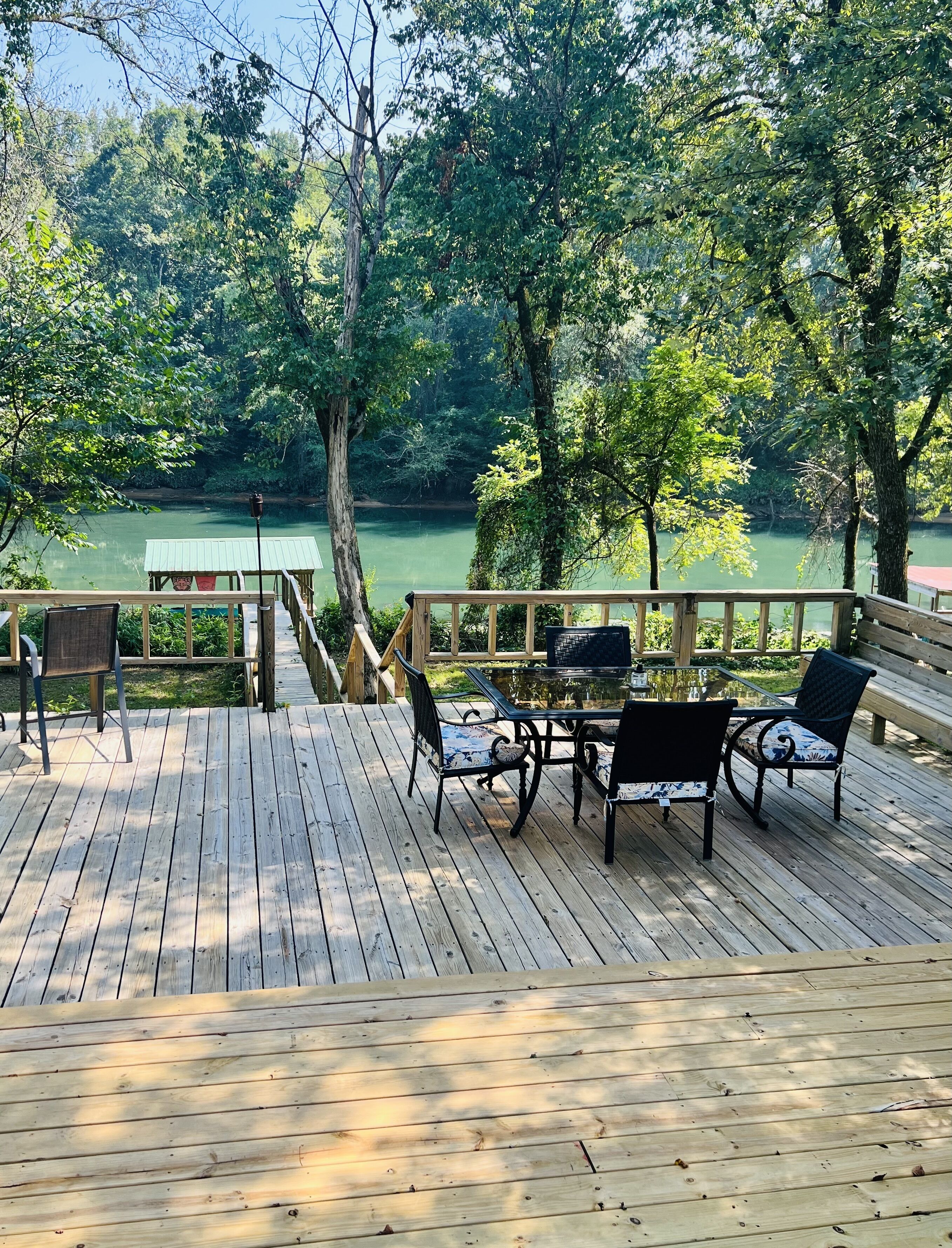 A wooden deck with outdoor furniture overlooking a calm body of water, surrounded by lush greenery.