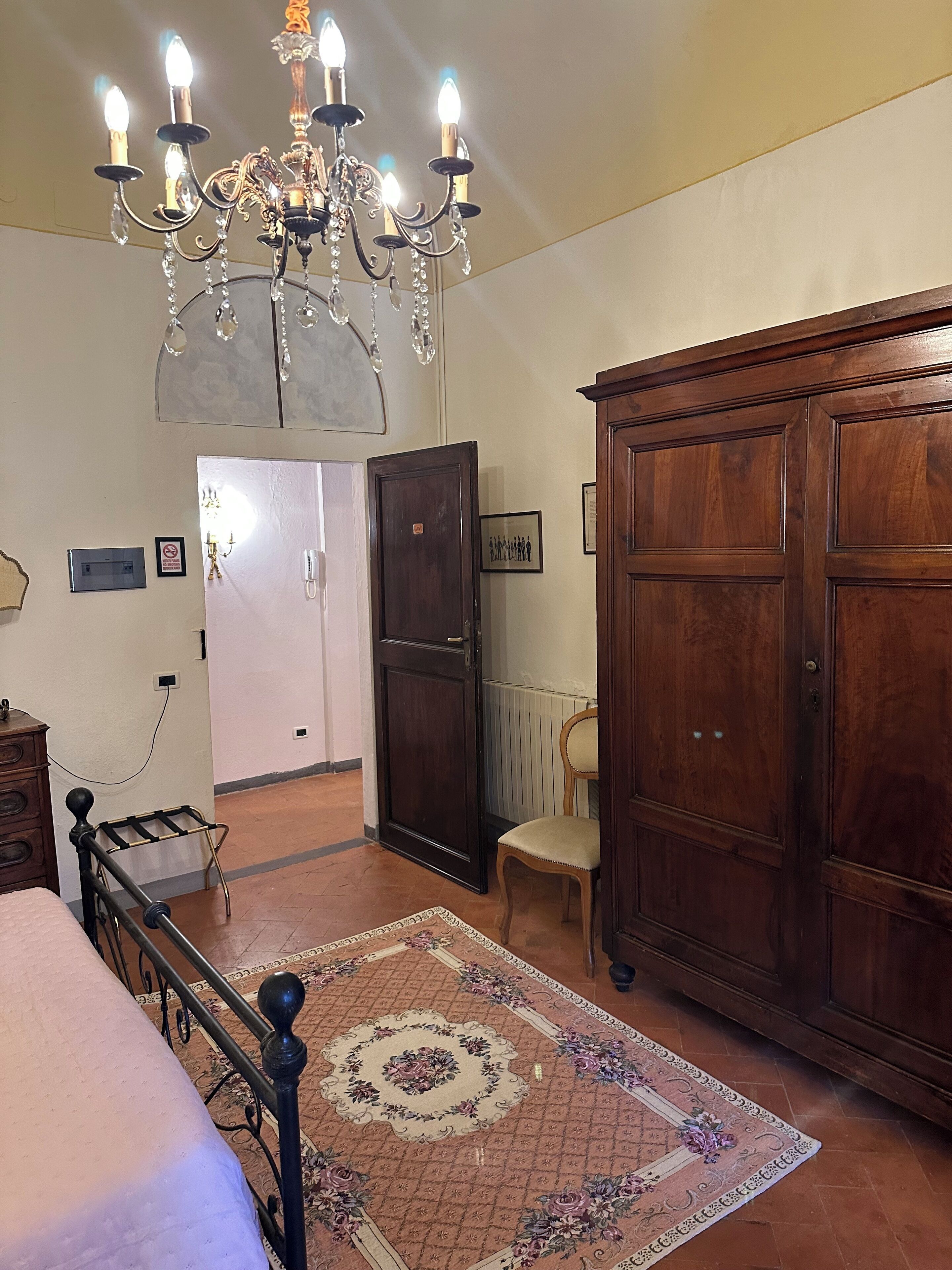 A bedroom with a bed, a wooden wardrobe, a chair, a chandelier, and a door leading to another room.
