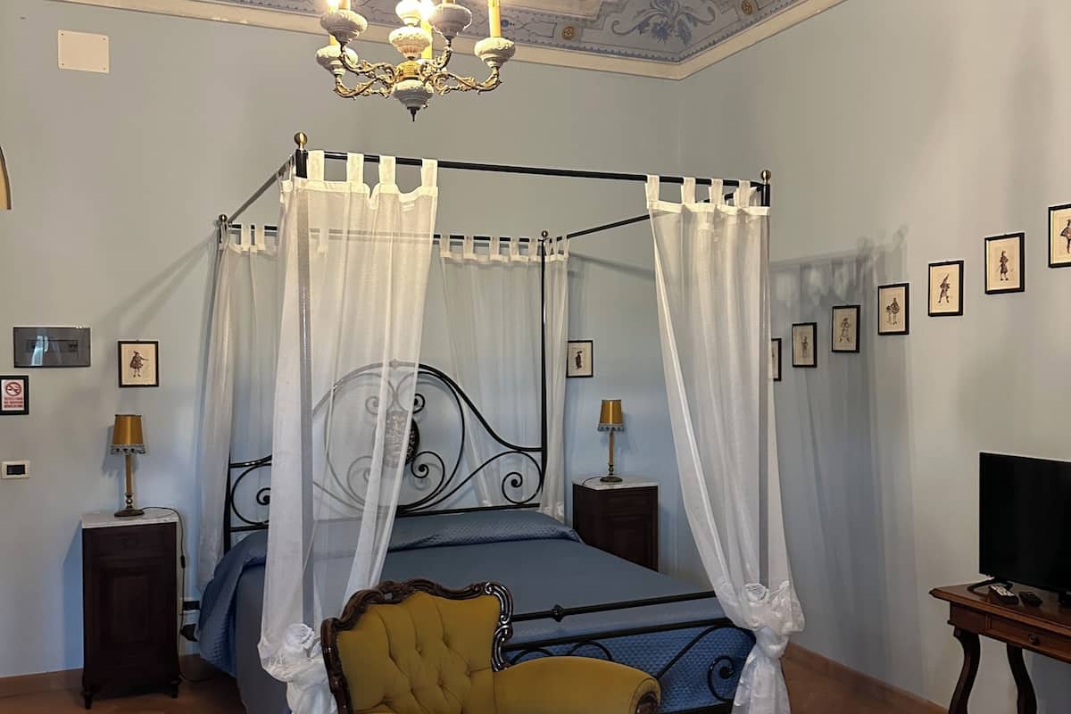 A bedroom with a four-poster bed, a yellow armchair, a chandelier, and a television on a wooden stand.