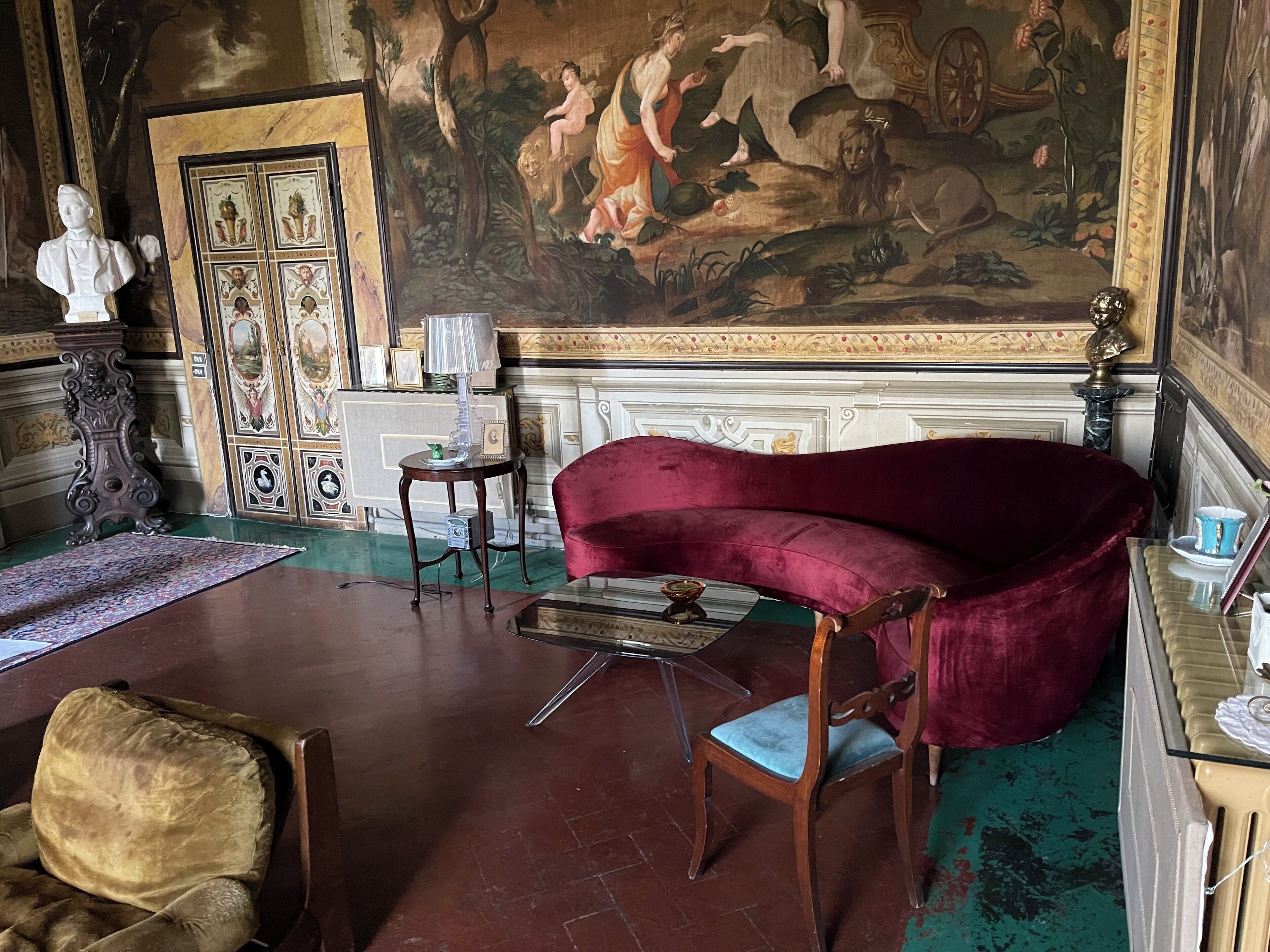 A room with a large classical painting, a red velvet sofa, a wooden chair, a small table with a lamp, and a bust sculpture.