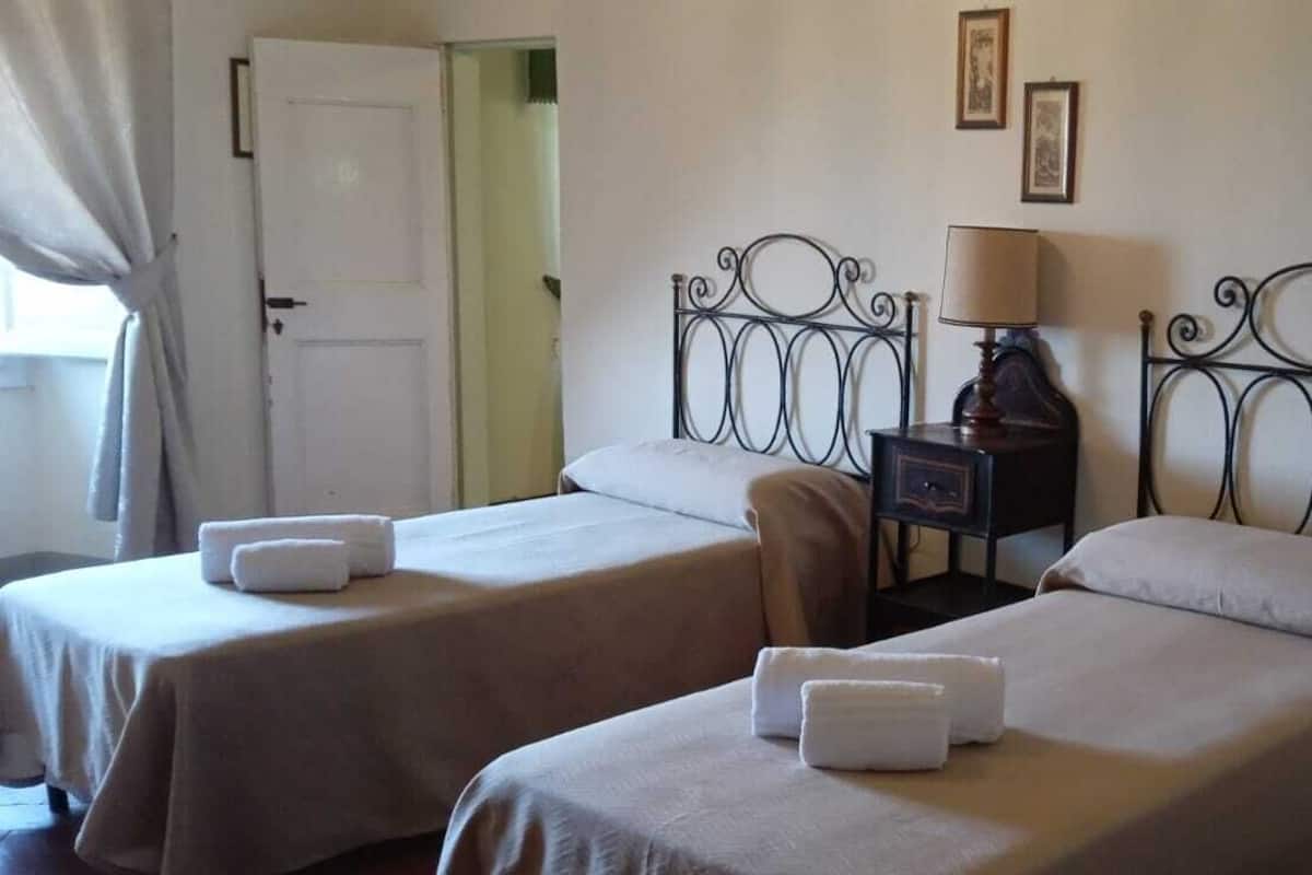 A hotel room with two single beds, each with a white bedspread and a towel. There is a nightstand with a lamp between the beds.