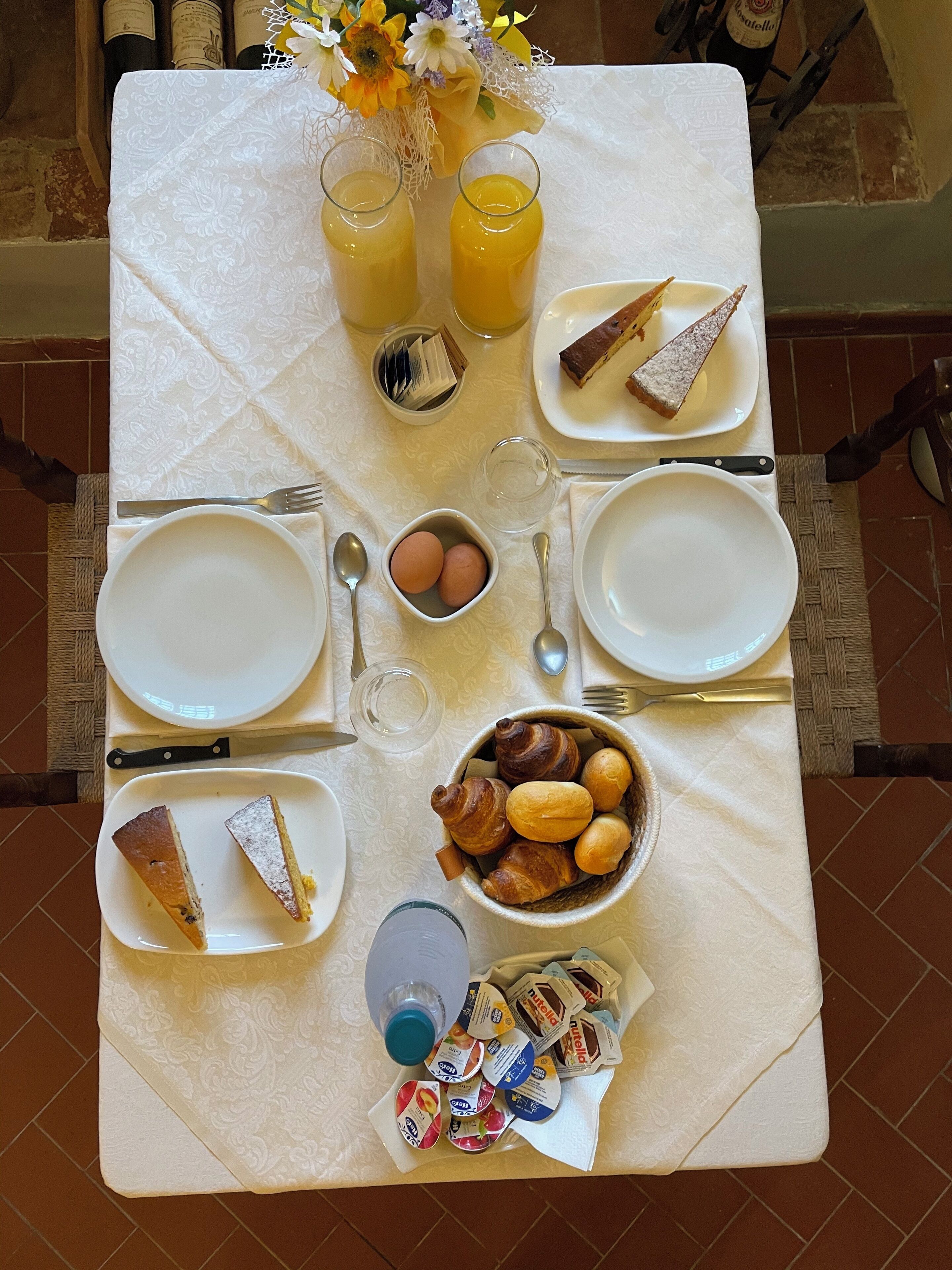 Mesa de café da manhã posta com pratos, talheres, copos e alimentos, incluindo pães e ovos.