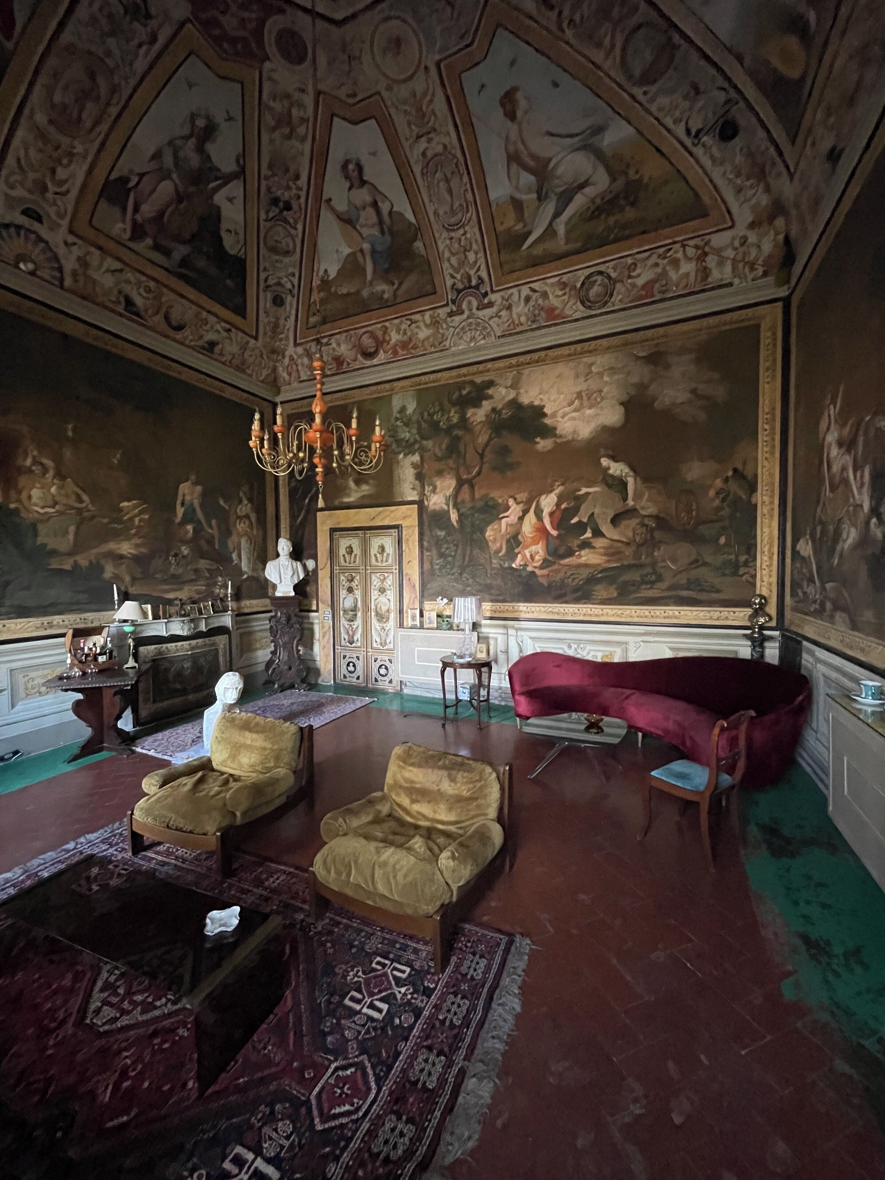 Ein Zimmer mit aufwendigen Deckenfresken, einem Chandelier und klassischen Möbeln, darunter ein rotes Samtsofa und Sessel.