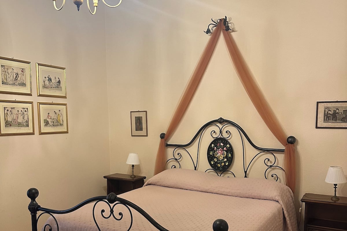 A bedroom with a bed, a chandelier, framed pictures on the wall, and a wooden floor.