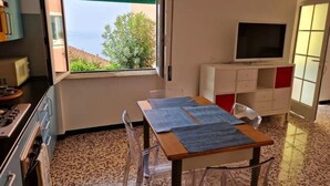 A small kitchen with a window, a table with placemats, a TV on a stand, and a refrigerator. - Panoramic garden on the sea, a corner of Liguria (Sant'Andrea di Rovereto)