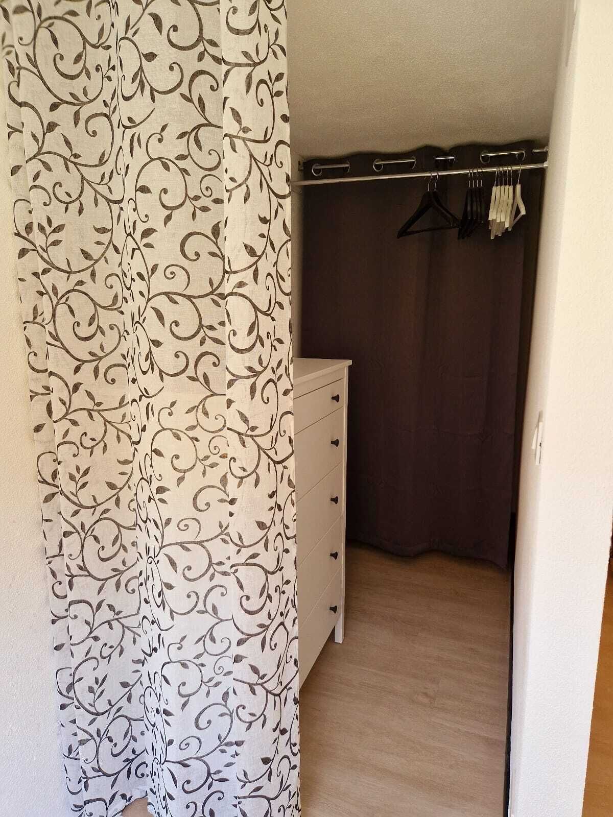 Ein Zimmer mit einem gemusterten Vorhang, einem weißen Schminktisch und einem Kleiderschrank mit Kleidung.