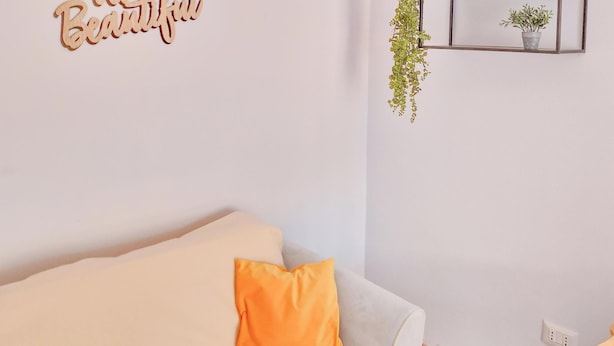 A room with a white wall, a wooden shelf with three mugs, a hanging light, and a cushion on a sofa.