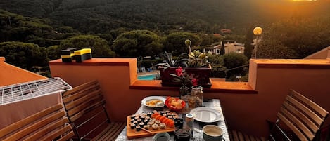 A rooftop dining area with a table set for a meal, surrounded by chairs, and overlooking a mountainous landscape.