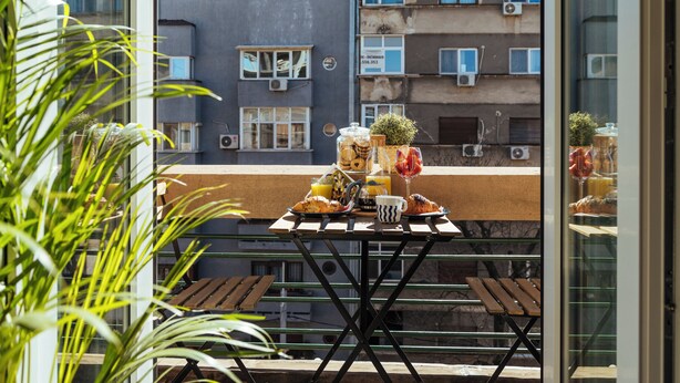 Un balcone con tavolo apparecchiato per la colazione, affacciato su un edificio con numerose finestre e balconi.