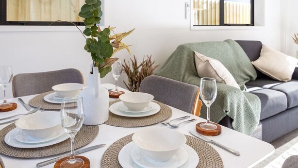 Un comedor con una mesa dispuesta para cuatro, incluyendo platos, tazones, cubiertos y vasos. Hay una planta en un jarrón blanco, y un sofá gris con cojines al fondo.