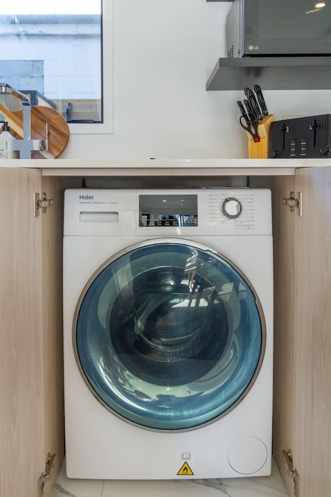 Una lavadora Haier blanca y compacta de carga frontal, con panel de control digital, ubicada en un área de cocina con armarios de madera y un microondas arriba.