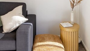 Una sala de estar moderna con un sofá gris, una mesita redonda de madera con un jarrón y objetos decorativos, y un ventanal con persianas.