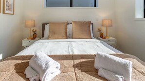 Una cama bien hecha con sábanas blancas y dos almohadas color beige, flanqueada por mesitas de noche con lámparas y jarrones decorativos.