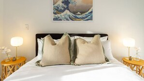 A neatly made bed with two pillows, a framed artwork above, and two bedside tables with lamps and flowers.