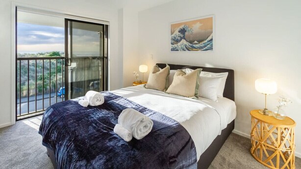 A hotel room with a bed, a balcony, a painting, and a side table with a lamp.